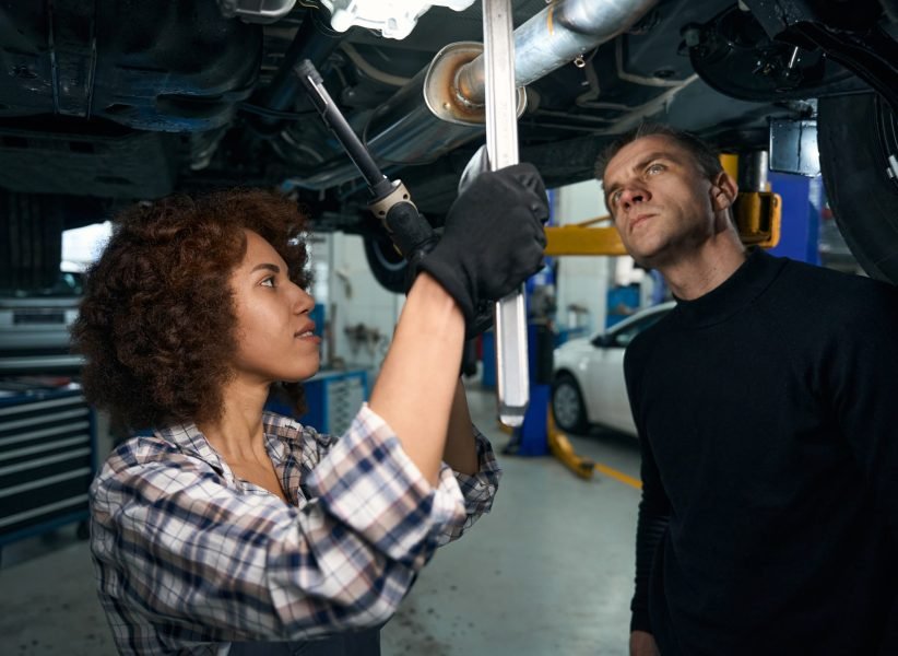 Young woman auto mechanic and a client inspect the car from below, the woman has a special lamp